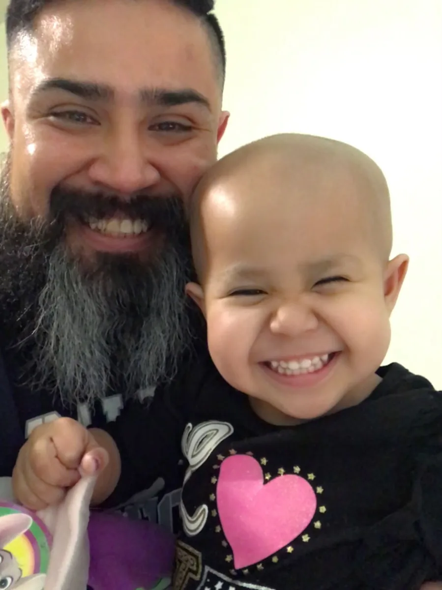 Joel Ojeda, with a full beard, smiling close beside his young daughter Mia, who is grinning with her eyes squeezed shut