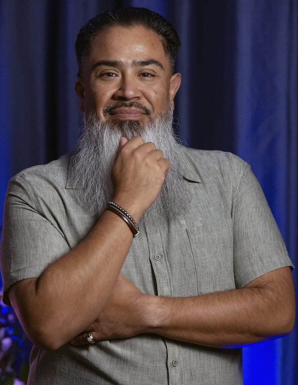Joel Ojeda, smiling, with one hand at his chin, in front of a blue curtain